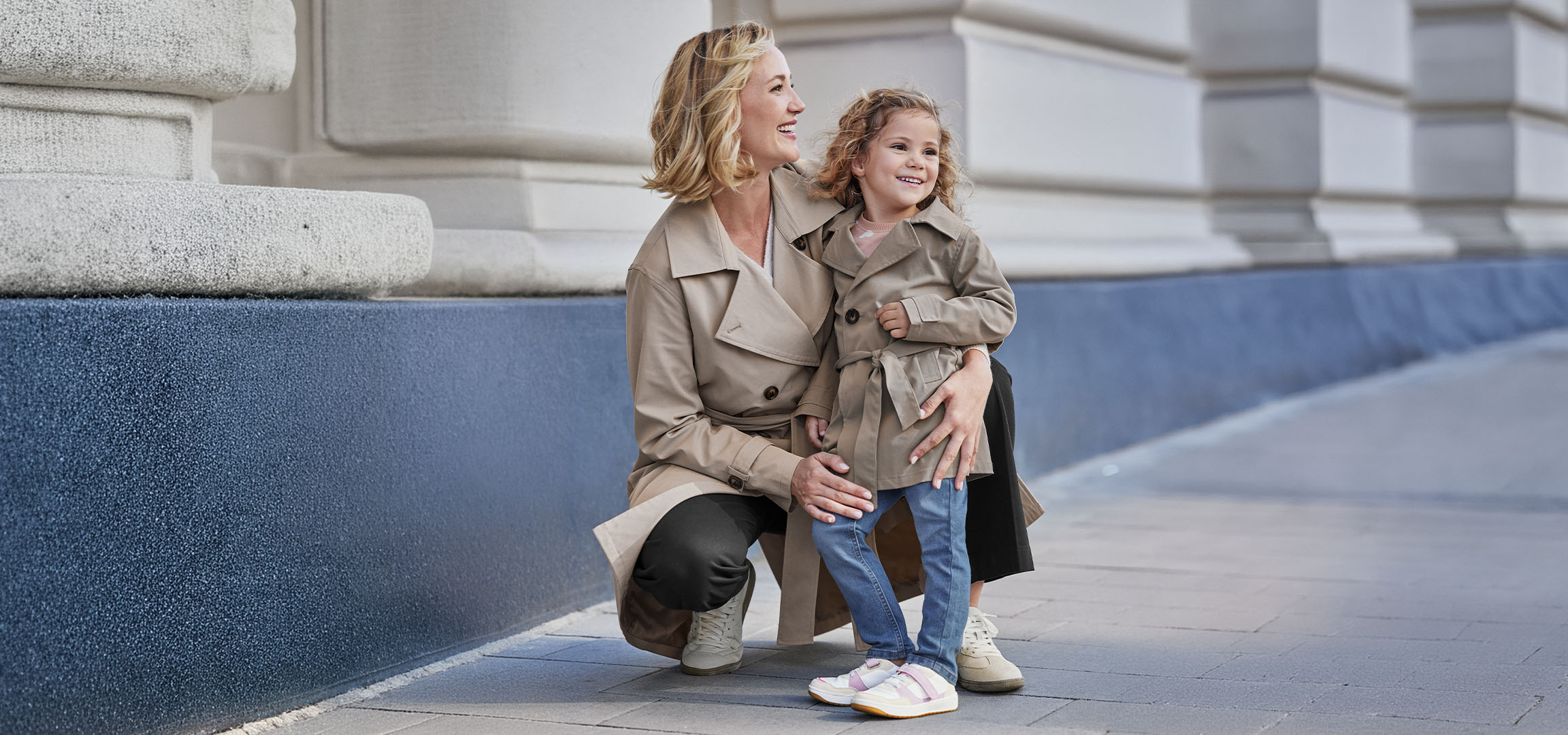 Twee vrouwen in beige trenchcoats, een volwassene en een kind, lachend buiten.