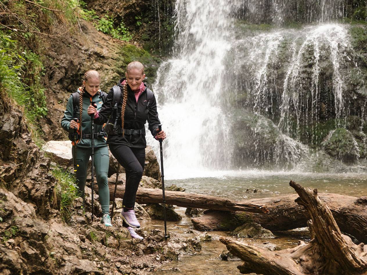 Twee vrouwen met wandelstokken lopen over een rotsachtig pad bij een waterval.