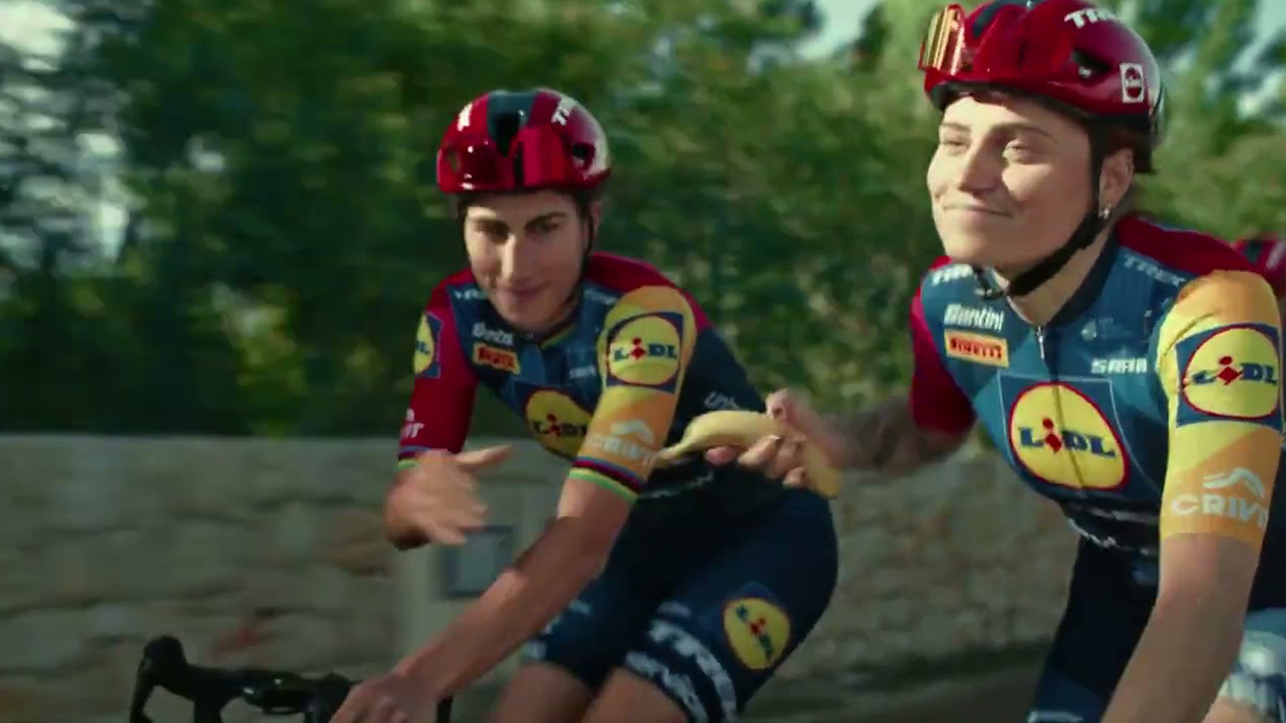 Twee fietsers in sportkleding, waarvan één een banaan aan de ander geeft.