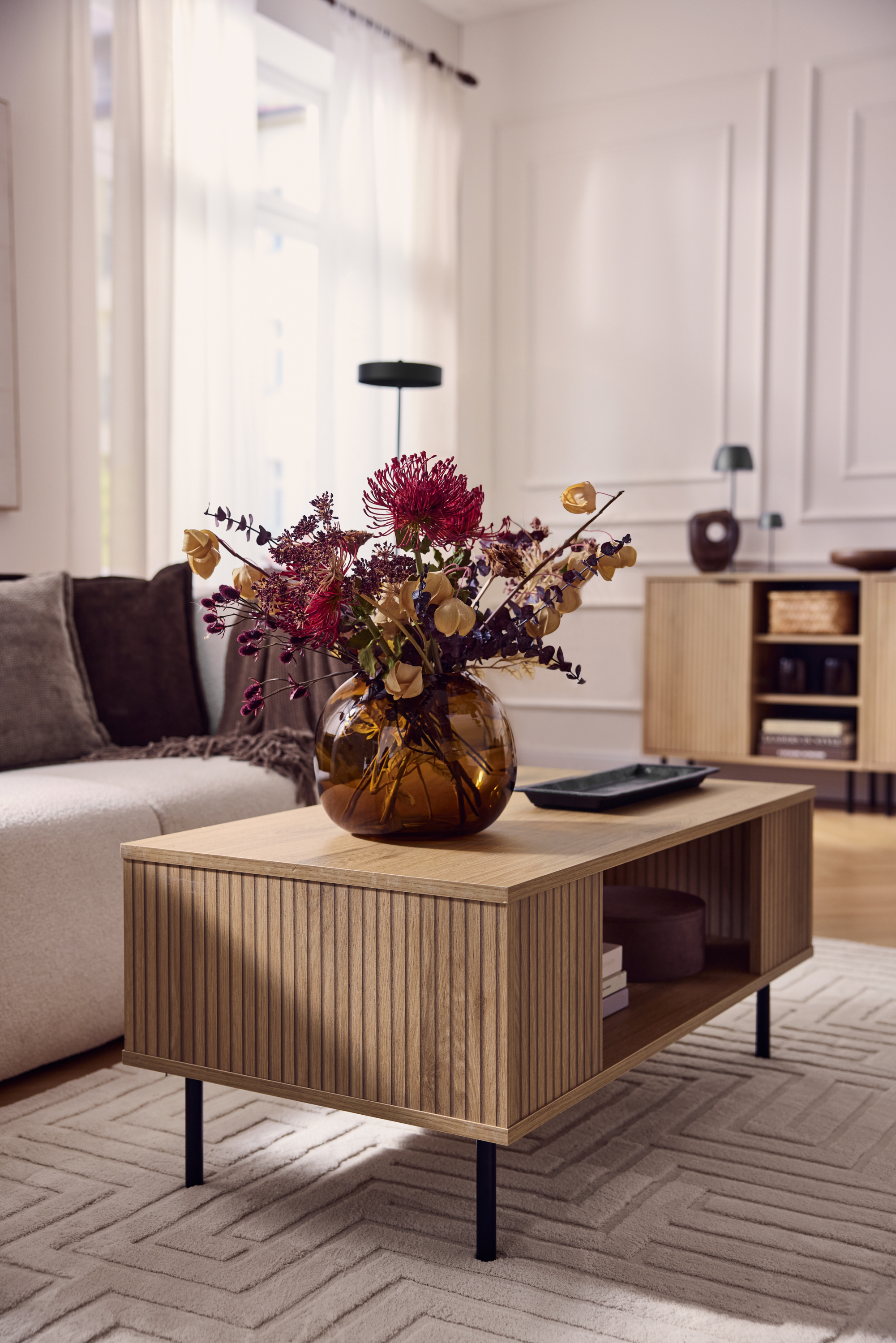 Houten salontafel met geribbelde voorkant, vaas met gedroogde bloemen, beige bank.